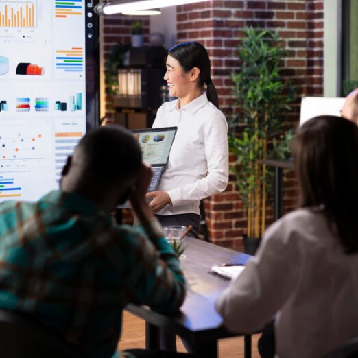 Female entrepreneur analyzes financial charts, cheerfully discussing marketing strategy with coworkers. Professional asian woman holding laptop, laughing during group discussing in brick wall office.
