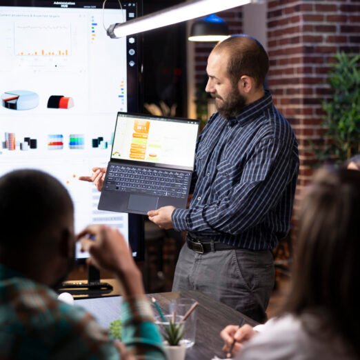 Caucasian male company advisor presents business analytics in laptop during late night group meeting. Diverse colleagues analyzing financial reports and brainstorming strategies in startup office.